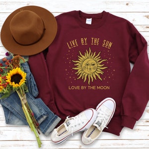 May include: Burgundy sweatshirt with a gold sun and moon graphic and the text "Live by the sun, love by the moon".