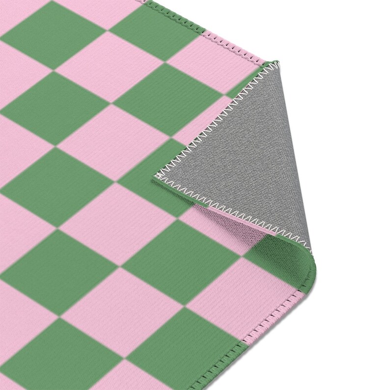 Puede incluir: Una alfombra a cuadros verde y rosa con un respaldo gris. La alfombra tiene un borde cosido.