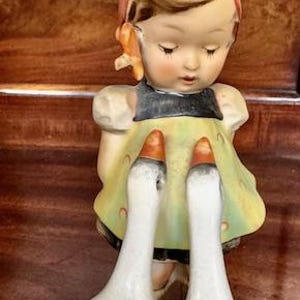 May include: A vintage porcelain figurine of a girl with two geese. The girl wears a pink headscarf, a black top, and a yellow dress. The geese are white with orange beaks. The figurine is on a round base.