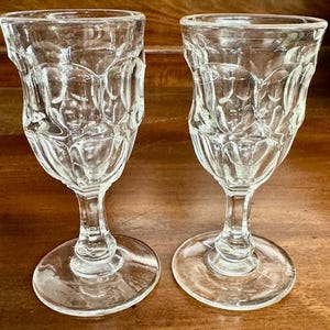 May include: Two clear, vintage-style stemmed glasses with a textured, repeating pattern. The glasses have a tulip-like shape and sit on round bases. The glass is clear, allowing light to pass through, and the design is visible.