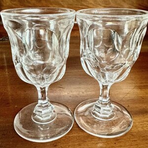 May include: Two clear, vintage-style stemmed glasses. Each glass features a decorative, embossed pattern on the bowl and a round base. The glasses are made of clear glass and are suitable for serving beverages.