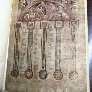 May include: A close-up of a page from an old book with a detailed illustration of a medieval manuscript. The illustration is in shades of brown, black, and white and features a series of columns with text and decorative elements. The text is in a medieval script.