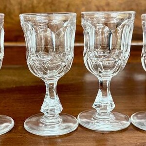 May include: Four clear, stemmed shot glasses with a decorative, cut-glass design. Each glass has a round base, a faceted stem, and a flared, patterned bowl. The glasses are arranged in a row on a wooden surface.