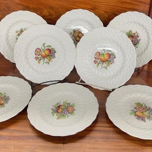 May include: A collection of white ceramic plates with a textured basketweave pattern. Each plate features a central fruit design with peaches, grapes, and other fruits. The plates are arranged on a wooden surface.