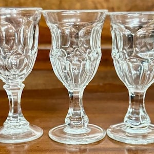 May include: Three clear, vintage-style cordial glasses with ornate, textured designs. Each glass features a flared bowl, a slender stem, and a circular base. The glasses are displayed on a wooden surface.