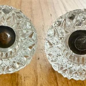 May include: Two round clear glass knobs featuring a faceted diamond-cut pattern across the surface. Each knob has a circular dark metallic center insert with a recessed hole, providing a contrast to the transparent, reflective glass exterior.