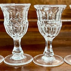May include: Four clear, stemmed glasses with ornate, textured designs. The glasses have a tulip-shaped bowl, a slender stem, and a round base. They are arranged on a dark wooden surface.