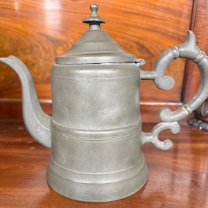 May include: A vintage pewter teapot with a hinged lid, curved spout, and ornate handle. The teapot has a cylindrical body with horizontal bands and a small finial on top. The metal has a matte, gray finish, and the design suggests an antique style.
