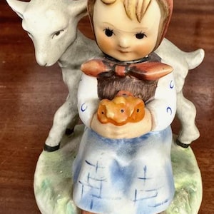 May include: A ceramic figurine of a young girl in a blue dress and red bonnet, holding a basket of bread. A lamb stands behind her. The base is green with the words "Good Friends".