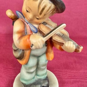 May include: A ceramic figurine of a young musician playing a violin. The figurine is wearing a dark hat, orange jacket, blue bow tie, and green pants. The figurine is standing on a round, off-white base.