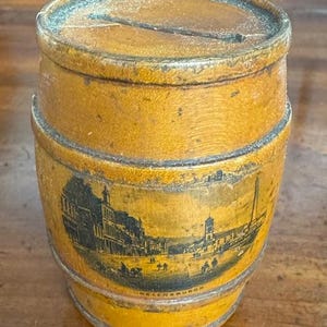 May include: A vintage wooden barrel-shaped money bank with a slot on top. The bank is painted yellow with black metal bands and a black and white image of a town scene. The word "HELENSBURGH" is printed on the bank.