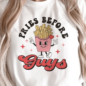 Puede incluir: Camiseta blanca con un gráfico de un contenedor de papas fritas de dibujos animados con el texto "Fries Before Guys" en rojo y negro.