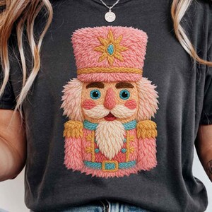 Pode incluir: T-shirt cinzento escuro com um desenho de quebra-nozes rosa e dourado. O quebra-nozes tem um chapéu, barba e uniforme rosa com detalhes dourados e azuis. A imagem inclui o texto "ONLY PNG 300DPI".