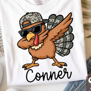May include: White t-shirt with a cartoon turkey wearing sunglasses and a camouflage hat, in a dabbing pose. The turkey is brown, with a red wattle and a yellow beak. The name "Conner" is printed below the turkey in black script.