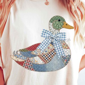 May include: Cream-colored t-shirt featuring a patchwork duck design. The duck has a green head, a yellow beak, and a blue gingham bow. The body is made of various fabric patterns, including floral and denim.