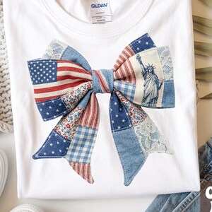 Puede incluir: Camiseta blanca con un gran diseño de lazo que incorpora la bandera estadounidense, la Estatua de la Libertad y detalles de patchwork de mezclilla. El lazo es el elemento central, con tonos rojos, blancos y azules. La camiseta está doblada sobre una superficie blanca.