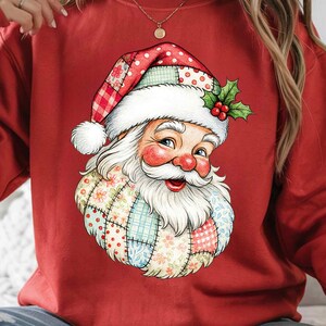May include: Red sweatshirt featuring a Santa Claus graphic. The Santa Claus illustration has a white beard, rosy cheeks, and a patchwork design on his hat and coat. The hat is red with white polka dots and a holly sprig.
