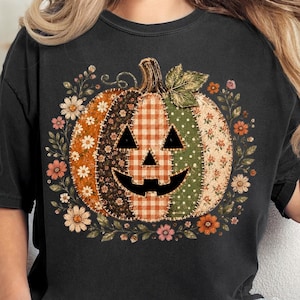 Könnte beinhalten: Ein schwarzes T-Shirt mit kurzen Ärmeln und einem großen Kürbis-Motiv. Der Kürbis besteht aus verschiedenen stoffartigen Mustern wie orangefarbenem Vichy-Karo, grünen Punkten und Blumenmustern und ist von einem Blumenkranz umgeben.