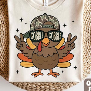 May include: Cream-colored t-shirt featuring a cartoon turkey wearing sunglasses and a camouflage hat. The sunglasses read "GOBBLE GOBBLE". The turkey is giving the peace sign with both hands. Black star accents are scattered around the turkey.