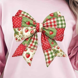 May include: Pink sweatshirt with a large bow design. The bow is a patchwork of red polka dots, green gingham, and strawberry-patterned fabric. The bow's centre is red and the overall design has a vintage, country aesthetic.