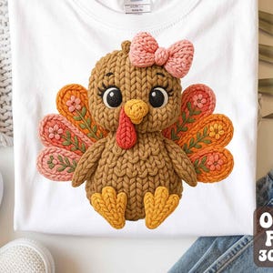 Puede incluir: Camiseta blanca con un diseño de pavo tejido. El pavo tiene un cuerpo marrón, un lazo rosa y plumas coloridas en tonos naranja, rosa y verde. La camiseta está doblada y expuesta sobre una superficie neutra.