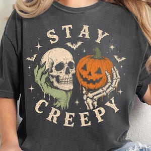 May include: A charcoal gray t-shirt featuring a graphic print with the text STAY CREEPY. The design includes a human skull held by a green hand and a jack-o'-lantern pumpkin held by a skeletal hand, surrounded by small stars and flying bats.