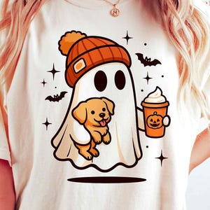 May include: Cream-colored t-shirt featuring a cartoon ghost wearing an orange beanie and holding a golden retriever puppy. The ghost also holds a pumpkin spice latte. Black bats and stars decorate the design, perfect for Halloween.