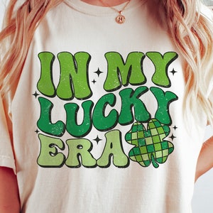 May include: Cream-colored t-shirt with the text "IN MY LUCKY ERA" in green, retro-style lettering. A green and white plaid shamrock is below the text. The shirt has a relaxed fit and is suitable for St. Patrick's Day.