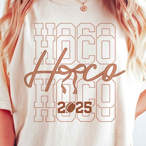 Könnte beinhalten: Cremefarbenes T-Shirt mit braunem Grafikdesign. Das Design zeigt den Text "HOCO" vertikal gestapelt, mit "Hoco" in Schreibschrift und einer Football-Grafik mit dem Jahr "2025".