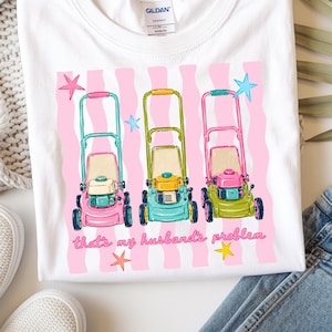 May include: White t-shirt featuring a graphic of three lawn mowers in pink, green, and blue, set against a pink striped background. The text reads "that's my husband's problem".