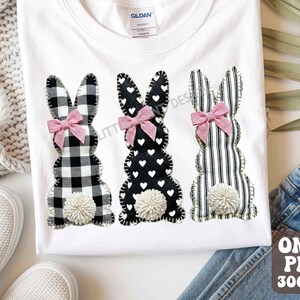 Puede incluir: Camiseta blanca con tres diseños de conejitos de Pascua aplicados. Cada conejito tiene un patrón diferente: cuadros blancos y negros, negro con corazones blancos y rayas blancas y negras. Cada uno tiene un lazo rosa y una cola de pompón blanca.