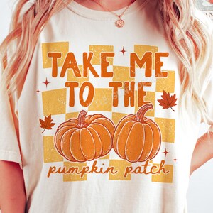 Può includere: Una t-shirt color crema con una grafica su sfondo a scacchi giallo e arancione. Il testo "TAKE ME TO THE pumpkin patch" è scritto in arancione e marrone. Due zucche arancioni e piccole foglie d'acero sono al centro del disegno.