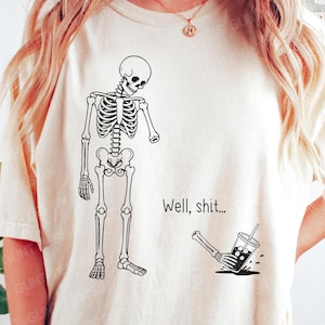 May include: A cream-colored t-shirt featuring a black line-art graphic of a skeleton missing one arm. The detached arm is shown on the ground holding a spilled iced coffee cup. The text "Well, shit..." is printed in black next to the skeleton.