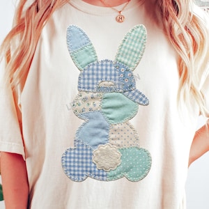 Pode incluir: T-shirt creme com um design de coelho em patchwork. O coelho é composto por vários tecidos estampados em azul e branco, incluindo xadrez, floral e bolinhas. O design está centrado na camisa, criando uma estética caprichosa e divertida.
