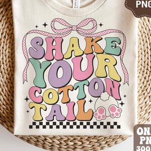 May include: A beige t-shirt with a colorful graphic that says "Shake Your Cotton Tail". The graphic features a pink bow, a bunny tail, and a checkered pattern.