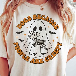 May include: A white t-shirt with a graphic design of a ghost holding a skeleton dog. The text around the graphic says "Dogs because people are creepy".