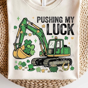 May include: Cream-colored t-shirt featuring a green excavator with shamrocks in its bucket and around the design. The text "PUSHING MY LUCK" is printed above the excavator. Additional St. Patrick's Day themed graphics include a pot of gold, a horseshoe, and a leprechaun hat.