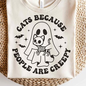 May include: A cream-coloured t-shirt with a Halloween graphic. The design shows a ghost holding a skeleton cat, with bats and stars, and the text "CATS BECAUSE PEOPLE ARE CREEPY" in a circular layout.