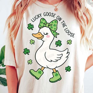 May include: Cream-colored t-shirt featuring a cartoon goose with a green bow, boots, and shamrocks. The text "LUCKY GOOSE ON THE LOOSE" is arched above the goose. The design is whimsical and festive, perfect for St. Patrick's Day.