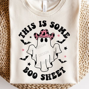May include: A white t-shirt with a ghost wearing a pink and black cowboy hat. The text "This is some boo sheet" is printed on the shirt.