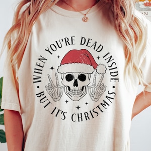 Pode incluir: Camiseta creme com um design gráfico circular. O design apresenta uma caveira usando um gorro de Papai Noel, fazendo o sinal de paz com mãos esqueléticas. O texto diz: "WHEN YOU'RE DEAD INSIDE BUT IT'S CHRISTMAS."