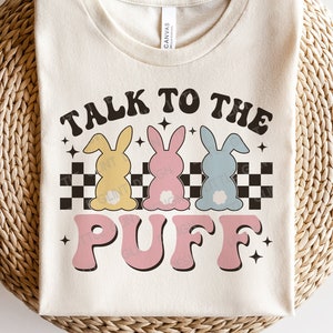 May include: A white t-shirt with the text "Talk to the Puff" in pink and black letters. Three cartoon bunnies in yellow, pink, and blue are behind the text. The bunnies are standing on a black and white checkered pattern.