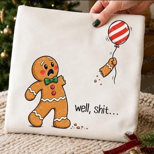 May include: A white sweatshirt featuring a cartoon gingerbread person with a shocked expression, a green bow tie, and two red buttons. One arm is detached, floating upward while holding a red and white striped balloon. The text reads well, shit...