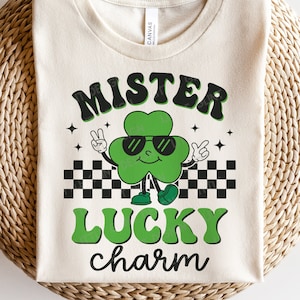 May include: A white t-shirt with a green four-leaf clover wearing sunglasses and a checkered pattern behind it. The text "Mister Lucky charm" is written in green and black letters.
