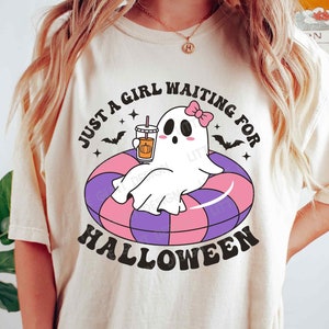 Może przedstawiać: Biały T-shirt z grafiką przedstawiającą ducha w r&oacute;żowej kokardce trzymającego latte z przyprawą dyniową w r&oacute;żowym kubku. Duch siedzi w fioletowo-r&oacute;żowym dmuchanym k&oacute;łku. Na koszulce nadrukowany jest czarny napis "Just a girl waiting for Halloween".