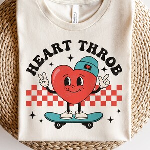 Op de afbeelding: Een wit T-shirt met de tekst "HEART THROB" in rode en blauwe letters. Een rood hart met een blauwe hoed en armen skateboarded op een geruit patroon.