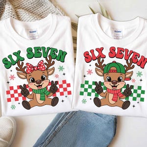 May include: Two white t-shirts with a Christmas theme. Each shirt features a cartoon reindeer with the text "SIX SEVEN". One reindeer wears a red bow, the other a green cap. Both have peace signs.