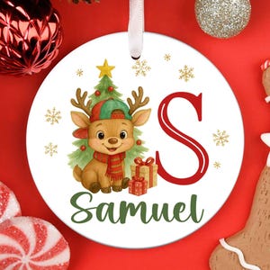 May include: A white Christmas ornament with a cartoon reindeer wearing a hat and scarf, a Christmas tree, and the letter "S". The name "Samuel" is written below the reindeer. Gold snowflakes and a star decorate the ornament.