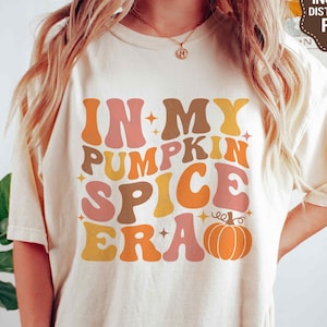 May include: A white t-shirt with the text "In My Pumpkin Spice Era" in a retro font with orange, yellow, pink, and brown colors. A small orange pumpkin with a green stem is included in the design.