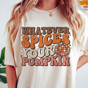 May include: A white t-shirt with the text "Whatever Spices Your Pumpkin" in a retro font. The text is in shades of orange, brown, and pink. A disco ball pumpkin is featured below the text.